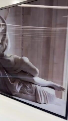 The video shows a close-up of a framed black and white photograph depicting a marble sculpture of two figures in a classical style. The sculpture shows one figure reclining while the other is standing, possibly in a moment of embrace or struggle. The camera then pans to the right along a glossy, white wall with three recessed lighting fixtures. The wall appears to be paneled with horizontal lines or grooves. Another framed black and white photograph appears on the wall, depicting a man.