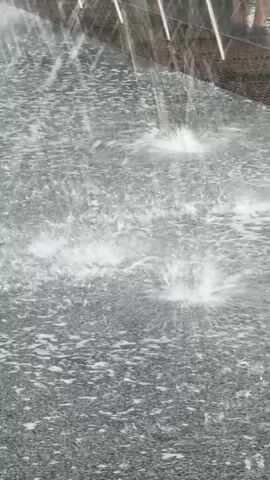 This media file captures a close-up view of a water feature, likely a fountain or a water play area. The primary subject is the water itself, with numerous droplets and splashes creating dynamic patterns on a textured, dark surface, possibly asphalt or stone paving. In the background, a row of thin, vertical structures, likely part of the fountain's mechanism or an adjacent architectural element, are visible. A person's legs and feet are partially visible at the top right, wearing sandals, suggesting a warm, possibly sunny day. The water appears to be spraying from multiple points, creating a lively and refreshing scene. The lighting suggests it could be daytime, with bright reflections on the water. The overall mood conveyed is one of playfulness and urban refreshment. There is no visible text in the media file. The location context of Piatykhatky, Ukraine, suggests this is an outdoor public space within that city.