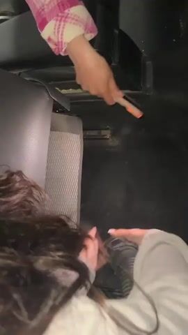 A woman in a grey sweater and another person in a pink and white checkered jacket are cleaning the floor of a car. The woman in the grey sweater uses the nozzle of a vacuum cleaner to suck up dirt while the other person uses a brightly colored marker to point out the spot to vacuum. The car has black carpet and a seat covered in grey fabric.