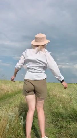 A blonde woman wearing a white button-down shirt, khaki shorts, a straw hat, and sunglasses poses in a grassy field. She is standing on a dirt path and moving her arms as if dancing. The sky is cloudy.
