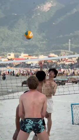 The video shows a group of young men playing beach volleyball. The volleyball is in the air, and one of the players is in the process of hitting it. The scene is set on a sandy beach with a volleyball net in the foreground. In the background, there are mountains and a beach populated with many people. The sky is clear, and it appears to be daytime. The players are all shirtless, wearing beachwear. The atmosphere seems casual and recreational.