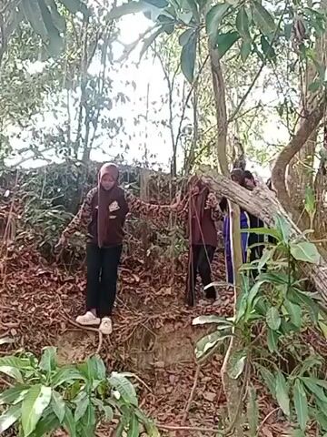 A group of young people are walking through a wooded area. Some are wearing headscarves and long clothing. They are on a path that appears to be eroding. The environment is dense with trees and foliage. In one shot, the group is near a body of water, possibly a river, with a small waterfall in the background. Another shot shows a young man looking towards the camera. The overall setting appears to be a natural, outdoor environment.
