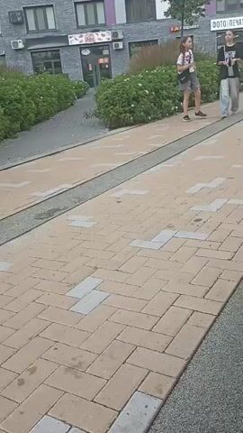 Three young girls walk along a paved walkway. The girl on the left wears a white t-shirt with a graphic design, grey shorts, and black sneakers. She carries a dark bag over her left shoulder. The middle girl is dressed in a black t-shirt, light-colored baggy pants, and dark sneakers. She holds a black phone in her right hand. The girl on the right wears a colorful t-shirt, pink shorts, and dark sneakers. She has a light pink crossbody bag. The walkway is paved with beige and grey bricks in a herringbone pattern. A strip of gravel runs alongside the walkway. In the background, there are buildings with shops, landscaping, and trees. The overall environment appears to be an outdoor public space.