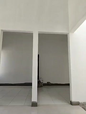The video pans across an interior space that appears to be under construction or newly built. The walls are white and unfinished in some areas, with some exposed concrete trim at the base. The floor is tiled with large, light-colored square tiles that reflect the light.

In the foreground, there is a large opening that leads to an outdoor area. This opening is framed by white pillars and a white horizontal beam. Through this opening, we see a construction site with piles of sand or dirt, some building materials, and a low-rise building with a brown and white facade. There is also a glimpse of a blue sky.

To the left of the main opening, there is another smaller opening, leading to a separate interior space, also with white walls and tiled floors.

The overall impression is one of a clean, unfinished, and open space with a view of an active construction area. There are no people visible in the video.