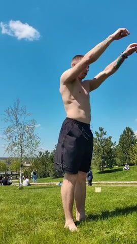 The video shows a shirtless man in black shorts performing a backflip in a park. The man stands on a grassy lawn under a blue sky with scattered clouds. He raises his arms, arches backward, and executes a backflip, landing back on his feet. In the background, there are trees, other people enjoying the park, and some buildings in the distance. The camera angle captures the entire action, and the lighting is bright due to the sunny day.