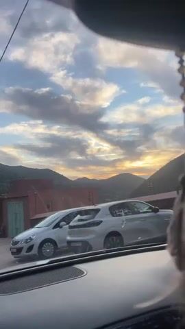 The video shows a view from inside a moving car, beginning with a partial view of a building and another car through the front windshield. The camera angle shifts upwards to capture the sky with scattered clouds and the setting sun on the horizon. The scene transitions to reveal a mountainous landscape, with green vegetation covering the slopes. The road ahead is visible, with a white car approaching in the distance. The environment suggests a rural or semi-rural area, likely in a mountainous region.