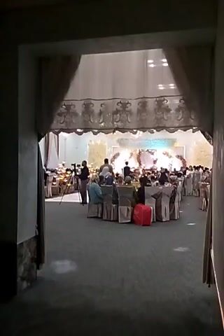 The video captures an indoor event hall viewed through an arched doorway. The hall is decorated with draped fabrics and lights, creating a festive ambiance. Several round tables are set up and covered with white cloths, each seating guests. In the foreground, a few people are visible, including a person holding a camera on a tripod. The middle ground shows more tables and guests, as well as what appears to be a stage area at the far end of the hall, adorned with decorative elements and lights. The overall scene suggests a wedding reception or a similar celebratory gathering.