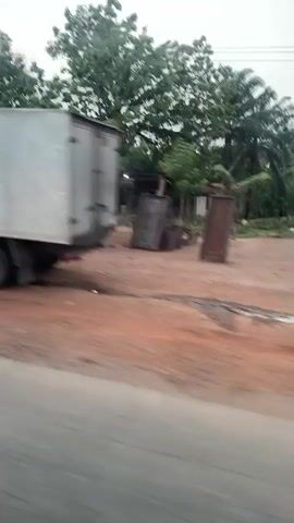 The video shows a car driving along a paved road in a rural area. At the beginning, a white box truck is seen connected to a smaller white truck. A person appears to be working in between the trucks. As the car continues to drive, it passes a gas station on the right side of the road, with yellow and white structures. Then, a man and woman are seen riding a motorcycle.  The area alongside the road has a lot of vegetation, and there are houses in the background. The sky is overcast.