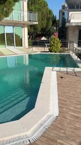 The video shows an outdoor swimming pool with crystal clear turquoise water. Steps lead into the shallow end of the pool. A pool cleaning device is submerged in the water. A hose is connected to the cleaner, extending out of the pool. A modern building with large glass windows and balconies is visible behind the pool, surrounded by lush green trees. The sky is clear and blue. The camera pans across the scene, showing different angles of the pool and the building.
