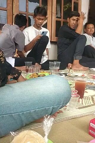 The video shows a group of people sitting on a mat in what appears to be a living room or dining area. There are several individuals, including men and what seems to be a child. They are engaged in a meal or gathering, with plates of food, drinks in cups, and a box of tissues visible on the mat. Some individuals are eating or talking, while others are looking at their phones or resting. The room has wooden doors and a window, and the lighting suggests it is daytime. The camera is positioned low to the ground, giving a close-up view of the scene.