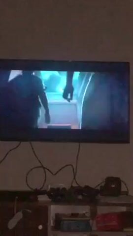 The video shows a television screen displaying a scene from a movie. The scene depicts two men walking down a hallway with orange and blue lighting. As the camera zooms in, the men are revealed to be wearing Roman-style clothing. Subtitles appear on the screen stating "Senator Gabinius shall join presently." The overall environment of the scene appears to be a Roman-style villa or palace. The room is dimly lit by torches on the walls. The camera angle is a mid-shot, focusing on the two men in the hallway.  Beneath the television screen, there are electronic devices with tangled wires on a shelf.