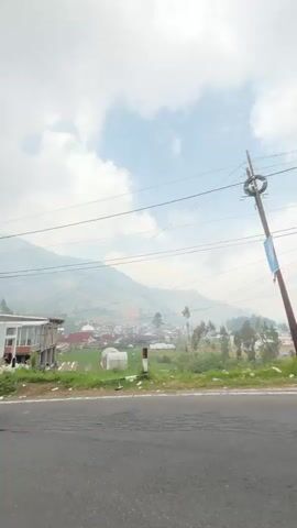 The video shows a roadside view of a village nestled in a valley. In the foreground, a paved road with cracks runs across the bottom of the frame, with a thin strip of grass and a few scattered items to the left. Beyond the grass, there are buildings and a mosque with a red roof and a white dome, and a wooden staircase. Several people are visible, including individuals standing near the mosque and a person on a red motorcycle. Further in the background, more buildings and houses are scattered across the green landscape, with rolling hills and mountains rising in the distance under a cloudy sky. Power lines stretch across the upper portion of the image.