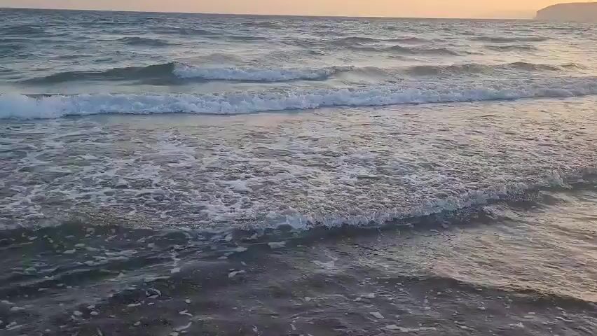 The video shows a close-up, eye-level view of ocean waves gently rolling onto a sandy shore. The water is a mix of blue and gray, with white foam cresting on the waves. The sunlight casts a warm glow on the horizon, creating a serene and peaceful atmosphere. The sandy beach is visible in the foreground, appearing dark and wet from the receding waves. No people or objects are visible in the frame. The camera remains static throughout the short clip.