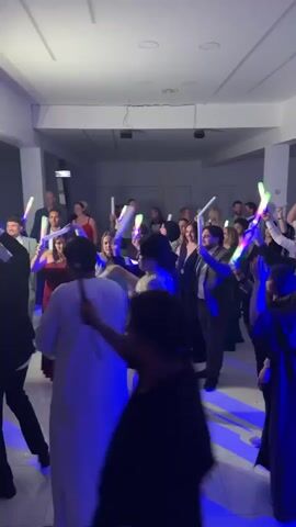 A wedding reception is in full swing, with many guests dancing under colorful party lights. The bride, in a long white sequined dress, leads the dancing with a lit-up wand in her right hand, followed by a man in traditional white Middle Eastern dress to her left, who also holds a lit-up wand. Other guests, dressed in suits and dresses, are similarly enjoying the music, raising their lit-up wands and dancing along. The room is well-lit with white walls and tile flooring, decorated with a few pillars. People are also taking pictures and videos of the event with their smartphones.
