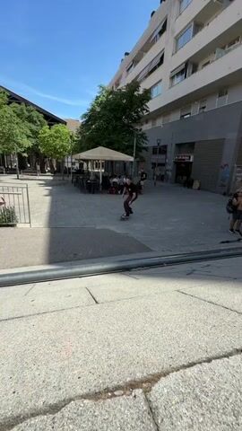 A young person wearing a black t-shirt and brown pants is skateboarding down a set of stairs outdoors on a sunny day. They approach the stairs, jump, and perform a trick in the air before landing on the pavement below and continuing to skate away. The skateboard has a green and yellow design. The background features buildings, trees, and some outdoor seating areas.