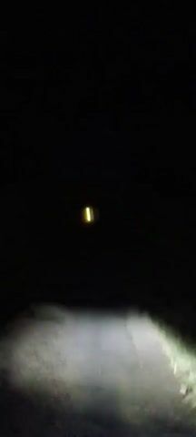 The video is filmed at night from the perspective of someone on a moving vehicle, likely a motorcycle, as indicated by the headlight beam and occasional view of handlebars and legs. The vehicle is traveling on a paved road that appears to be in poor condition, with visible cracks and uneven surfaces. The road is flanked by vegetation, which is illuminated by the headlights. The surroundings are dark, with trees and other natural elements visible in the periphery. In the distance, the headlights of other vehicles can be seen, and at one point, another motorcycle passes by. The audio contains spoken words in a language that is not English, likely conversational. The overall scene depicts nighttime travel on a rural or unlit road.