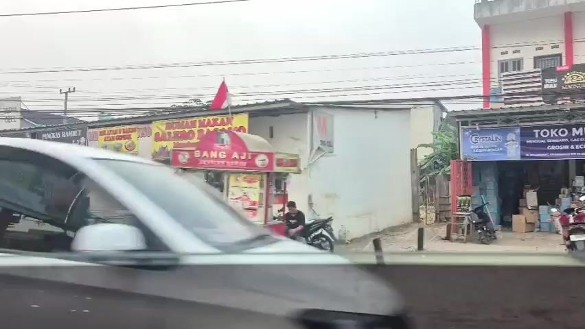 The video shows a street scene in what appears to be Southeast Asia. There are several small businesses lining the street, including a restaurant, a barber shop, and a food stall. Several motorcycles and a car are parked on the street, and a few people are visible walking and sitting outside the shops. An ambulance is parked in front of one of the buildings. The weather appears to be overcast. The video is shot from inside a moving vehicle, giving a blurred, impressionistic view of the surroundings.