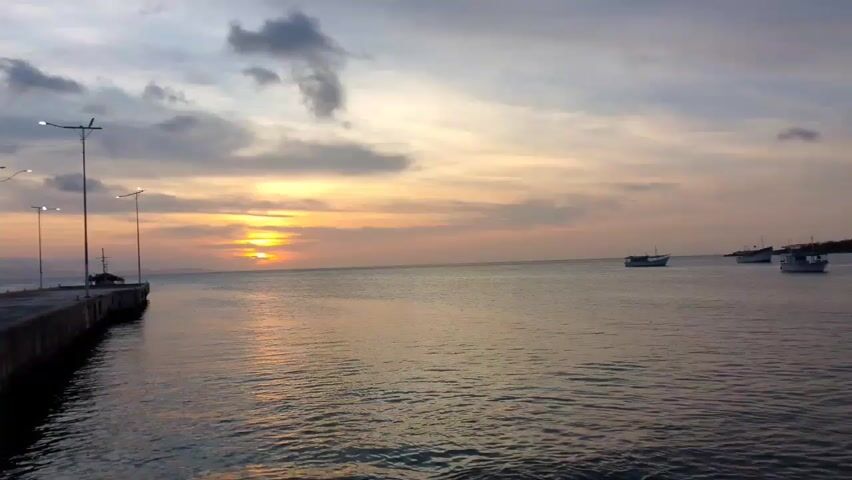 The video shows a sunset over a calm sea. A pier with street lights extends into the water from the left side of the frame. A person walks along the pier. Several boats are anchored in the water in the distance. The sky is a mix of orange, yellow, gray, and blue, with the sun partially obscured by clouds. The water reflects the colors of the sky. A mountain range is visible in the background on the right side of the frame. A voice speaks in the background.