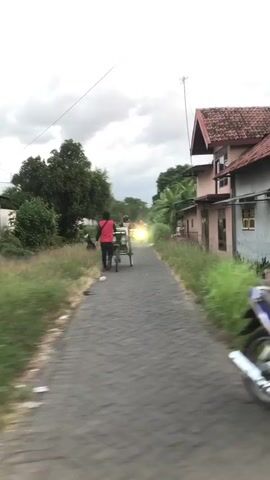 The video captures a narrow paved road with houses lining the right side and overgrown vegetation on the left. In the foreground on the right, a portion of a motorcycle is visible, suggesting the video is being filmed from a moving motorcycle. A person in a red shirt is walking away from the camera down the road, pulling a cart. Another person, whose clothing is indistinct, is seated on the cart. Further down the road, a bright light, possibly from a vehicle, illuminates the scene. The sky is overcast with clouds.