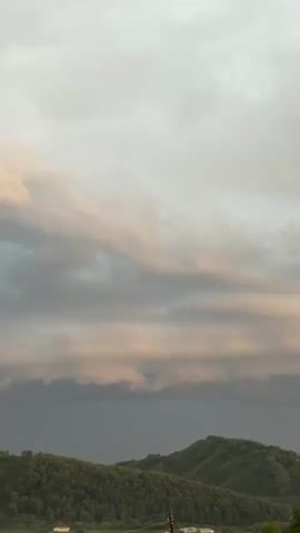 This is a vertical video capturing a landscape scene. The top portion of the frame is dominated by the sky, filled with a variety of clouds. The clouds are textured and appear to be in motion, with some areas of lighter, possibly sunlit, clouds in the upper part of the frame, transitioning to darker, more ominous clouds lower down. Below the clouds, a rolling landscape of green hills covered in dense trees stretches across the middle ground. The hills appear to be covered in lush vegetation. In the lower portion of the frame, a distant view of a rural area is visible, with hints of houses and possibly power lines. The lighting suggests it might be late afternoon or early evening, with soft, diffused light illuminating the scene. There are no people or distinct objects in the foreground, focusing the viewer's attention on the natural elements of the sky and the landscape. The overall impression is one of a serene yet potentially dramatic natural environment, possibly on the cusp of changing weather due to the prominent clouds.