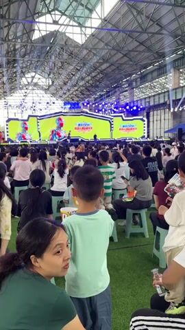 A wide shot captures an indoor event with a stage at the far end, illuminated by bright blue lights and featuring a large yellow screen displaying graphics, including a robot and Chinese text. A crowd of people, mostly children and adults, are seated on low plastic stools on a green artificial turf floor, facing the stage. The camera is positioned behind some of the audience members, focusing on their backs and heads. The event appears to be a performance or presentation, with a speaker on the stage. The overall atmosphere suggests a public gathering or festival.