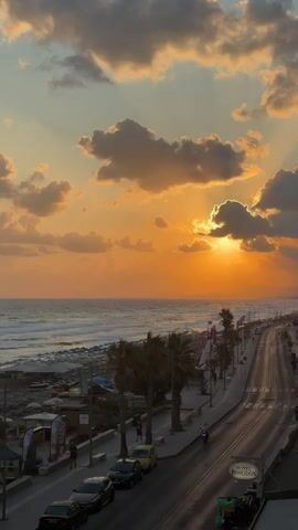 The video shows a sunset over a coastal town. The sun is setting over the ocean, casting a warm orange glow on the clouds and the water. The waves are crashing on the shore, and there are palm trees lining the street. Cars are driving on the road, and there are some people walking on the sidewalk. The overall scene is peaceful and beautiful.