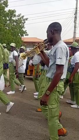 The media file shows a street parade in Dutse, Nigeria. The primary subjects are a group of young people, likely participants in a parade or celebration, dressed in matching green uniforms. Several individuals are playing musical instruments, including brass instruments like trumpets and percussion instruments such as drums. The parade appears to be moving along a paved street, with buildings and trees visible in the background.

The event taking place is a parade, characterized by the organized movement of people and the presence of musical performers. The participants are in uniform, suggesting a formal or organized group, possibly a school band, a youth organization, or a civic group. Their focused expressions and the act of playing music indicate a performance or procession.

Notable details include the time of day, which appears to be daytime with visible sunlight, suggesting it's not early morning or late evening. The weather seems to be fair, as participants are outdoors without heavy protective clothing. The presence of buildings, including what looks like a commercial establishment with signage, places the scene in an urban or semi-urban environment. Some individuals are carrying bags, and the overall atmosphere seems lively and engaged.

Visible text includes "SOUND" on the t-shirt of one of the individuals, and a banner that reads "ENFORCEMENT" and "OSSA STATE DRUG CON" followed by "THEME: THE WORLD DRUG ISSUES, NEW PERSPECTIVES, PERSISTING ISSUES, NEW APPROACHES, INNOVATIVE RESPONSE." This text clearly indicates that the parade is related to drug enforcement and awareness. The location context provided confirms it is Dutse, Nigeria.