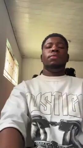 A medium shot captures a young, dark-skinned man wearing a white, long-sleeved shirt with a "JUSTICE" graphic featuring an image of handcuffs. He is positioned indoors, facing forward and looking around as if in thought. The room is simply furnished with a window on the left side, a light fixture on the ceiling, and a small part of a chair visible behind him.