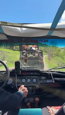 The camera is mounted in the front cab of an open-top jeep, following another jeep on a dirt road. The surrounding environment is rural, with grassy hills and trees lining the road. The road has patches of dirt and gravel. In the lead jeep, several people can be seen, including a woman in the passenger seat filming with her phone. The jeep behind is painted in army green and has an Armenian license plate. The interior of the lead jeep is visible, with gauges and dials on the dashboard. A phone is mounted to the dashboard. The sky is blue with some clouds. The steering wheel is held by a driver. A rear-view mirror shows the people in the jeep behind. A hanging car freshener is visible in the mirror frame.