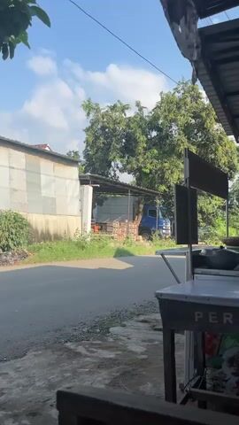 The video shows a daytime shot of a street scene, apparently taken from under the shelter of a building or structure with a corrugated metal roof. To the left is a low, silver-sided building and to the right is a food cart that has "PER" printed on the side of it.  Across the street, under an open-sided shelter, sits a large blue truck and a stacked set of red bricks. The sky is visible and is blue with some puffy white clouds. The road is gray asphalt.