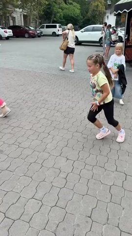 This media file shows a young girl performing a cartwheel on a paved walkway in Inkerman, Ukraine. The setting appears to be a public area, possibly a promenade or park, with trees and buildings in the background. Several other people are walking in the vicinity, some of whom are looking at the girl. The girl is wearing a light-colored t-shirt with a floral print, black leggings, white socks, and pink Crocs. She has her hair in a ponytail.

The girl initiates the cartwheel from a lunging position, quickly transitioning into the acrobatic move. Her body is fully extended as she moves through the motion. She lands back on her feet, appearing to be in control and with a smile on her face, suggesting confidence and enjoyment in her activity. The other individuals in the background are dressed in casual attire. The lighting suggests it is daytime, likely afternoon, with overcast conditions. The weather appears to be mild. No visible text is present on the girl's clothing or in the immediate surroundings. The overall atmosphere seems relaxed and casual.