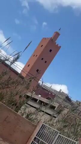 The video shows a tall, square minaret made of red brick against a blue sky with some clouds. The minaret has a few small windows on each side and a decorative top. The camera is angled upwards from ground level, looking past some bushes and buildings. The lower buildings are a mix of brick and concrete.