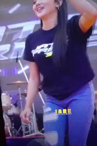 The video shows a young woman with long dark hair wearing a black t-shirt with a logo and ripped blue jeans. She is on a stage with a blurry background featuring stage lights and equipment. She is smiling and appears to be dancing or performing. There is another person in the background, a young man wearing a light blue shirt, who is also on the stage. A watermark "JARE ENTERTAINMENT" is visible on the woman's jeans.