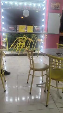 The video shows the interior of a restaurant with yellow chairs and tables. The chairs are arranged around a few tables, and some high chairs are placed near a counter. The counter is illuminated by a string of lights and has menus displayed behind it. The floor is tiled and reflects the lights, giving a shiny appearance. One person's leg and foot, wearing black shoes, are visible on the left side of the frame. The overall atmosphere appears casual and welcoming.