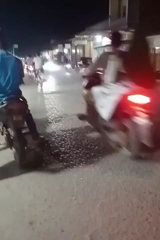 This video captures a street scene at night in Kruengmanyang, Indonesia. The primary subjects are several people on motorcycles, indicated by their presence and the illumination from their headlights and taillights. The setting appears to be a street lined with businesses, likely shops or stalls, as suggested by the structures and the goods displayed under artificial lighting.

The activity is centered around the movement of the motorcycles, with one vehicle in the foreground partially obscuring the view of others further down the street. The wet surface of the road, reflecting the lights, suggests recent rain or damp conditions. The overall atmosphere is that of a bustling night market or commercial area.

Notable details include the bright headlights of approaching vehicles, casting long shadows across the wet asphalt, and the red glow of taillights from vehicles moving away. The time of day is clearly night, given the darkness and the reliance on artificial lighting. The location cues, like the building structures and the overall ambiance, are consistent with a Southeast Asian urban or semi-urban environment. There is no discernible emotional tone conveyed due to the focus on the activity and the distance of the subjects.

Visible text includes "FOTO COPY" on one of the shopfronts, indicating a photo copying service is available. Another sign appears to read "OTO FIX", possibly related to automotive services.