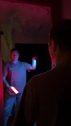 A person stands in front of a mirror, holding two illuminated LED light tubes, one red and one blue. The person's reflection in the mirror also holds the light tubes. The lighting in the room is dim and colored by the light tubes, with red and blue hues cast on the walls and ceiling. The person is wearing a t-shirt and jeans. The mirror is slightly cracked in the upper right corner.