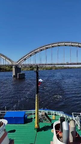 The view from the deck of a boat heading toward a bridge on a sunny day. The bridge is a through arch bridge, supported by concrete pillars. The boat is moving along a river or wide body of water. The deck of the boat is green and has some equipment on it. The sky is clear and blue.  The wind is blowing, ruffling the flag on the boat.