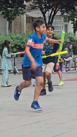A young boy in a blue and orange shirt and dark shorts runs towards the left of the frame holding a green foam baton. He is in a paved outdoor area with trees and buildings in the background. A woman in a white tank top and denim shorts walks away from the camera in the background. Other children and adults are also visible in the background, and a stroller is present. The scene appears to be a park or public square.
