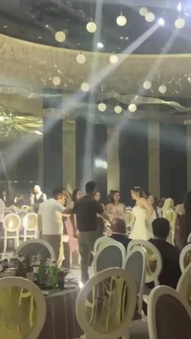 The video shows the inside of a large, fancy event hall. There are many people in the room, mostly dressed in formal attire. The hall is decorated with hanging lights, some resembling white globes. There are also tables with chairs around them, suggesting a dinner or reception setting. Spotlights move around the room, creating a dynamic lighting effect.  The camera moves a bit, trying to capture different angles of the scene and people.