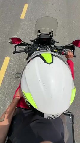 A person wearing a white and neon green helmet, a red long-sleeved shirt, and white shorts is seen riding a black adventure motorcycle on a winding asphalt road. The view is from above the rider's head, looking forward. The road features a distinct solid yellow center line and is surrounded by green hills and trees.

Initially, the rider's right hand is on the handlebars. Later, the rider is shown holding a smartphone in their left hand while riding, with the phone screen appearing dark. The weather transitions from bright and sunny with some clouds to predominantly cloudy.

As the motorcycle navigates the curves, simple, utilitarian buildings come into view. These structures include a two-story concrete building with a metal railing and a single-story building with a corrugated roof, situated amidst green vegetation. A solar-powered street light and a concrete trough are visible near these buildings. Red and white striped barriers line parts of the roadside.

The video captures a journey through a rural or mountainous area, characteristic of Minh Sơn Commune, Vietnam.