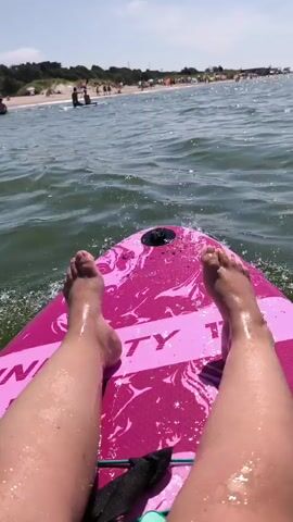 The video shows a first-person perspective of someone sitting on a pink paddleboard in the water. Their legs and feet are visible in the foreground, with the paddleboard’s branding, "INFINITY," clearly marked. The water is green and slightly choppy. In the background, a sandy beach is lined with trees and some structures. Several people are visible on the beach and in the water, some of whom appear to be on paddleboards or surfboards. The sky is clear and blue. The scene appears to be a sunny day at the beach.