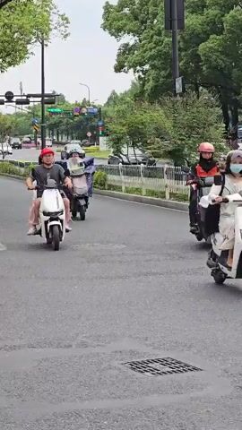 The video shows a street scene with several people riding electric scooters. In the foreground, a man wearing a red hat and a dark shirt is riding a white scooter with a woman wearing a pink outfit as a passenger. They are on the right side of the road. Behind them, another person on a scooter is visible, also covered with a blue tarp. Further down the road, a person on a scooter covered in a light-colored blanket is approaching. To the left, a yellow scooter is parked, with a red bag leaning against it. A crosswalk is visible in the lower left corner. The environment appears to be an urban or suburban area with trees and traffic lights. The weather seems to be overcast.