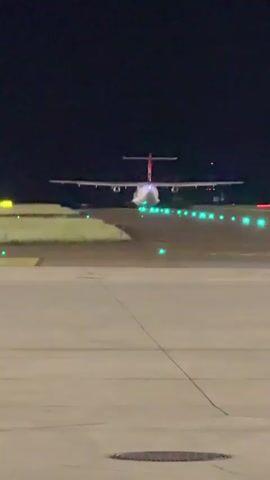 The video shows an airplane on a runway at night. The plane is facing away from the camera, and its engines appear to be active, indicated by the glow of the exhaust. The runway is illuminated by green lights, which are spaced along the sides. The sky is dark, suggesting it is nighttime. The camera is positioned on the tarmac, looking towards the runway.