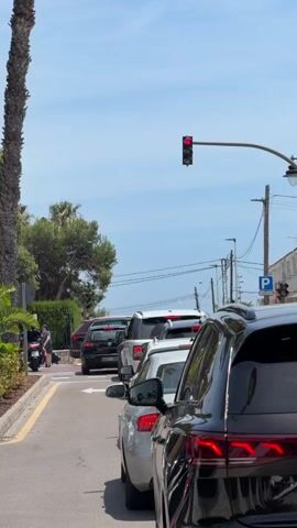 The video captures an outdoor scene on a sunny day, showing a line of vehicles stopped in traffic on a paved road. In the immediate foreground on the right, the rear of a black SUV is visible. Ahead of it, a silver sedan, a white SUV, a dark brown SUV, and a black hatchback are also stopped. An overhead traffic light displays a red signal, while a ground-level traffic light to the left shows a yellow signal. On the left side of the road, a sidewalk runs parallel to the street, bordered by green foliage including palm trees and hedges. A person on a white and red scooter is parked on the sidewalk on the left. Utility poles with wires are visible against the clear blue sky. A blue parking sign is affixed to a pole on the right side of the road.