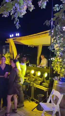 The video shows a nighttime outdoor party or event. In the first frame, a group of people are seen dancing and socializing under purple and yellow lighting. There are trees and a structure with yellow awnings in the background. The second frame shifts to a larger gathering of people dancing in an indoor or covered outdoor space with stone arches overhead and blue lights strung across the ceiling. The third frame shows a similar scene with many people dancing under purple and pink lights. The overall impression is of a lively social gathering with music and dancing.