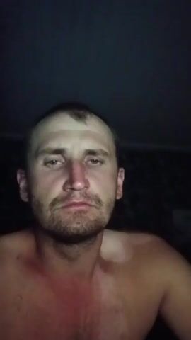 The video shows a close-up of a shirtless man with fair skin, short brown hair, and a beard. The man has a prominent forehead and green eyes. The lighting is dim, with the man's face illuminated from the front, creating shadows on his face and chest. His skin appears slightly red on his chest and neck, suggesting a possible sunburn or irritation. The background is dark and indistinct, with a slight blur that suggests movement or a low-resolution camera. The man's facial expressions change throughout the short clip, shifting from a neutral gaze to a slightly furrowed brow, and finally to wide, startled eyes before returning to a neutral expression. The overall impression is that of a selfie taken in low light conditions, possibly as part of a series of expressions.