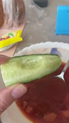 A close-up shot shows a person's hand holding a slice of cucumber over a bowl of red soup. The cucumber slice is green on the outside and light green with visible seeds on the inside. The soup appears to have small chunks of food in it. A spoon is visible in the soup bowl. In the background, a yellow packet and a brown loaf of bread are blurry. The scene is well-lit.