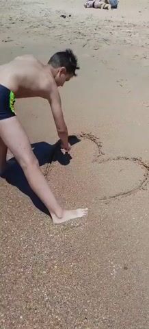 A young person wearing dark swim trunks with a green and yellow stripe is bent over on a sandy beach, drawing a heart in the sand with their finger. The sun is casting a shadow of the person onto the sand to their left. The background is a sandy beach with the ocean out of frame.
