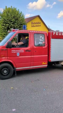 A red fire truck with the word "FEUERWEHR" and the number "112" visible is driving down a street. A person is driving the fire truck. The fire truck has a silver roll-up door on the back. There is a yellow building with "Buttstädter Volksbäckerei" written on it in the background. There are trees and a cloudy sky in the background.