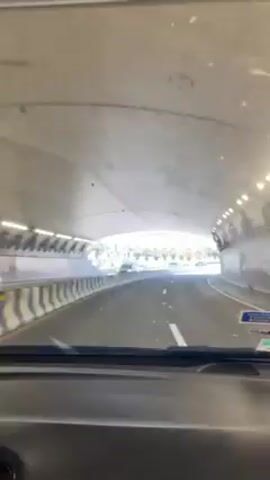 The video shows a car driving through a tunnel. The tunnel is well-lit and appears to be made of concrete. The car exits the tunnel and is now on a highway with palm trees and mountains in the background. There are other cars on the road. The sky is clear and blue. The day is sunny.